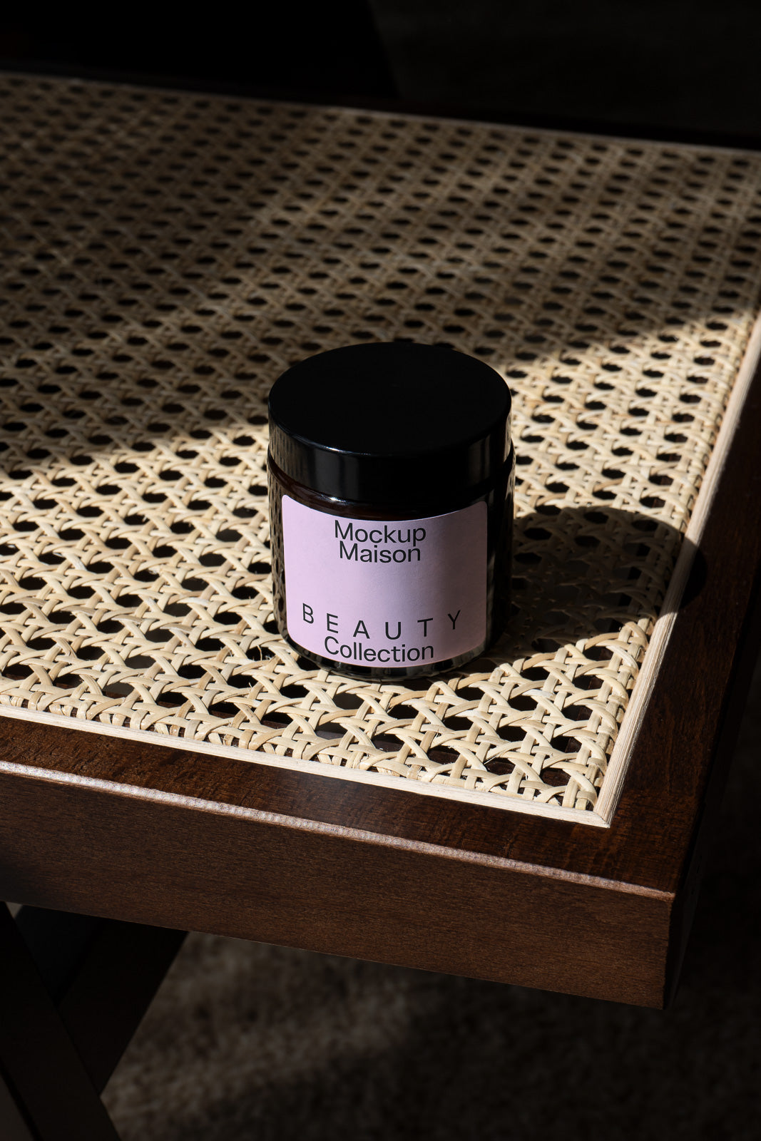 PSD mockup of a dark jar labeled "Mockup Maison Beauty Collection" placed on a woven wicker table surface.