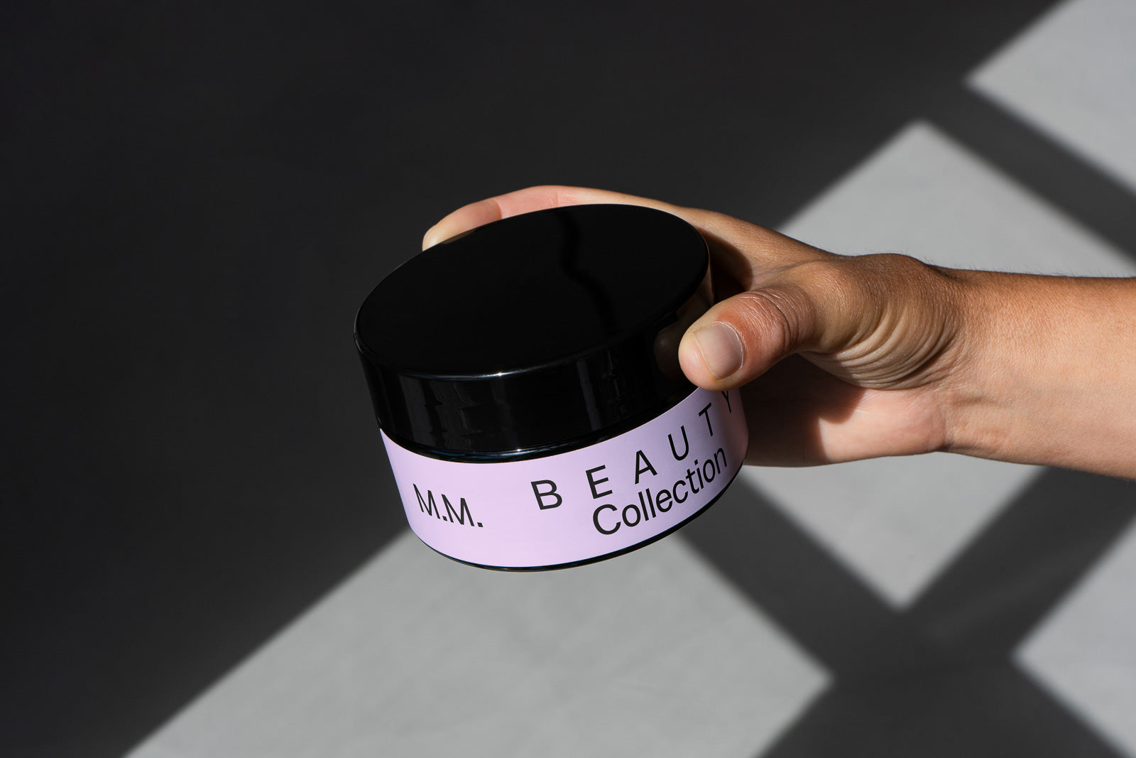 PSD mockup of a hand holding a beauty product jar labeled "M.M. Beauty Collection" with a black lid and lavender label.