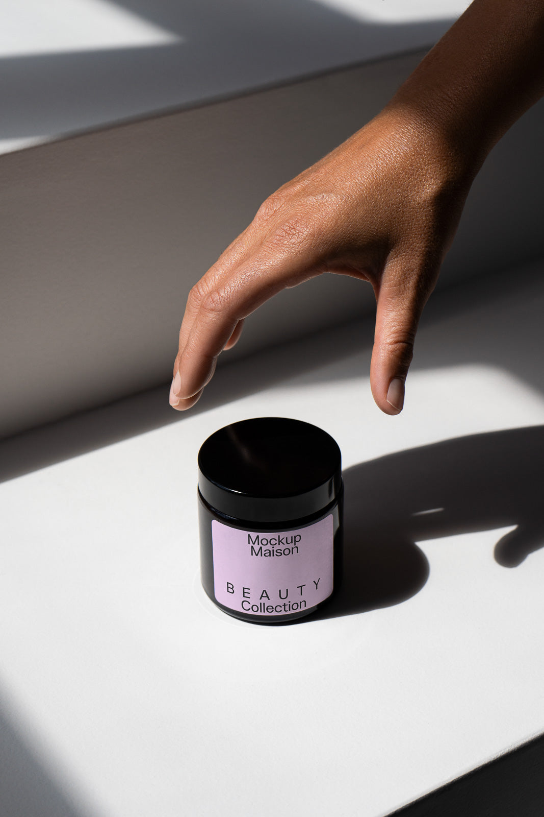 PSD mockup of a person’s hand reaching toward a black cosmetic jar labeled "Mockup Maison Beauty Collection" on a white surface, casting a shadow.