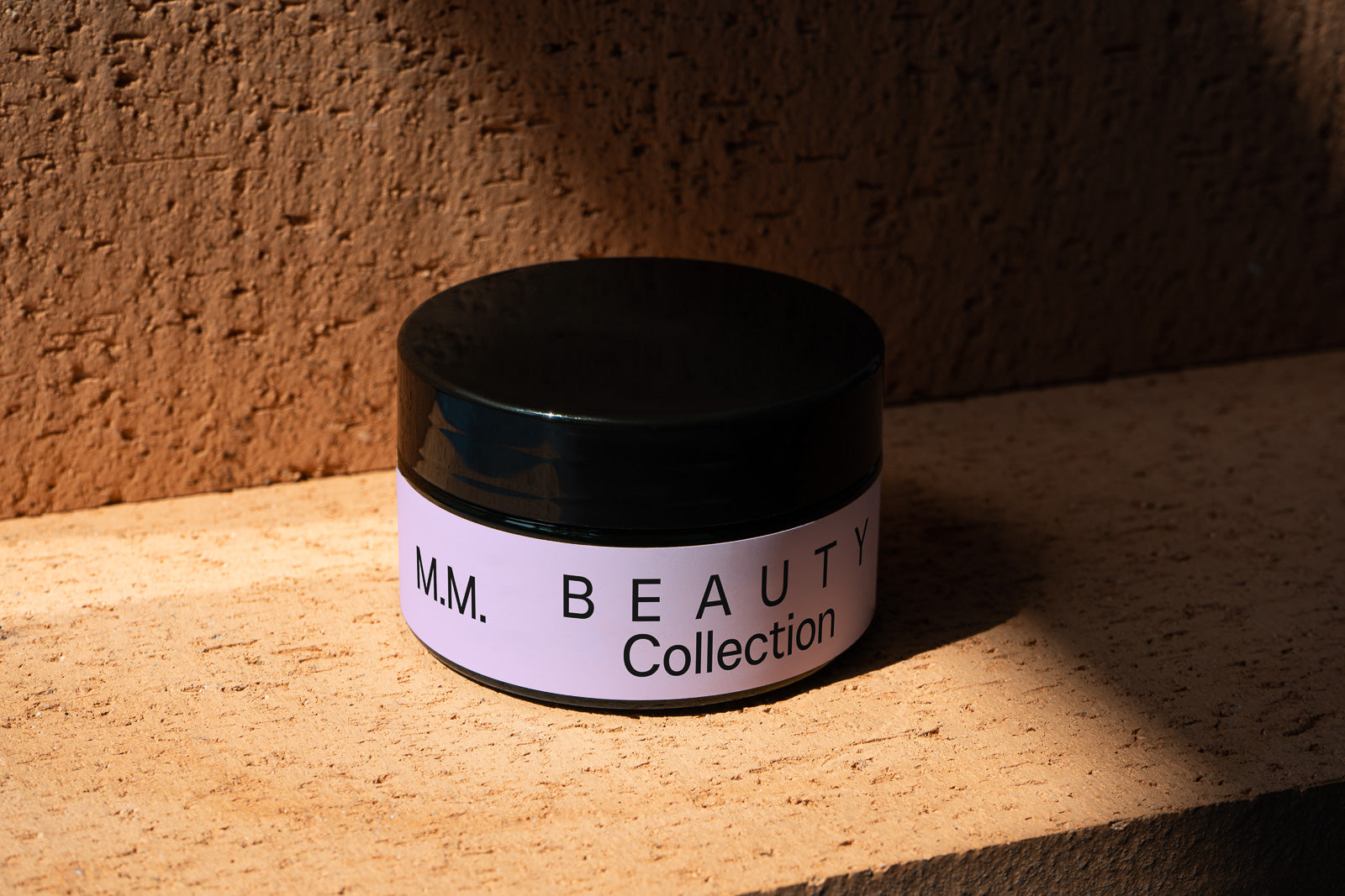 PSD mockup of a cosmetic jar labeled "M.M. Beauty Collection" with a black lid, set against a textured, earthy brown background, casting a soft shadow.