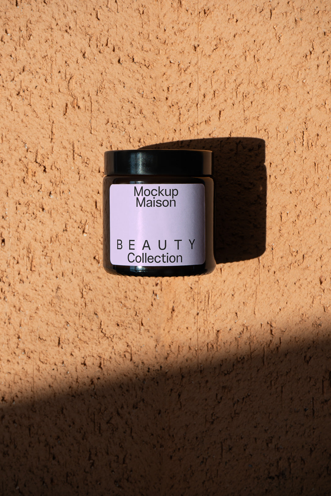 PSD mockup of a cosmetic jar labeled "Mockup Maison Beauty Collection" against a textured, sunlit wall with shadows.