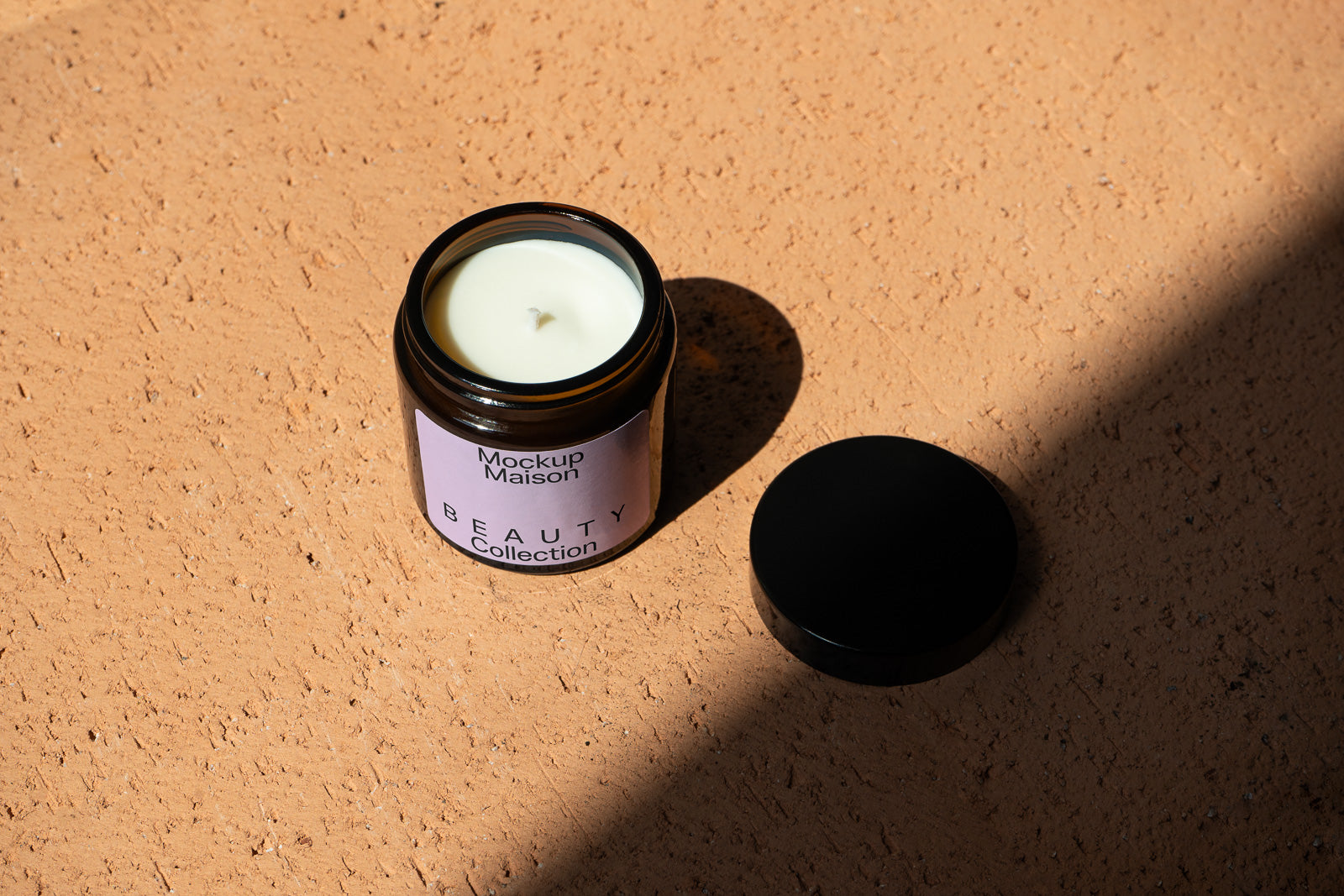 PSD mockup of a candle jar labeled "Mockup Maison Beauty Collection" on a textured surface with a shadow, lid off, and warm lighting.