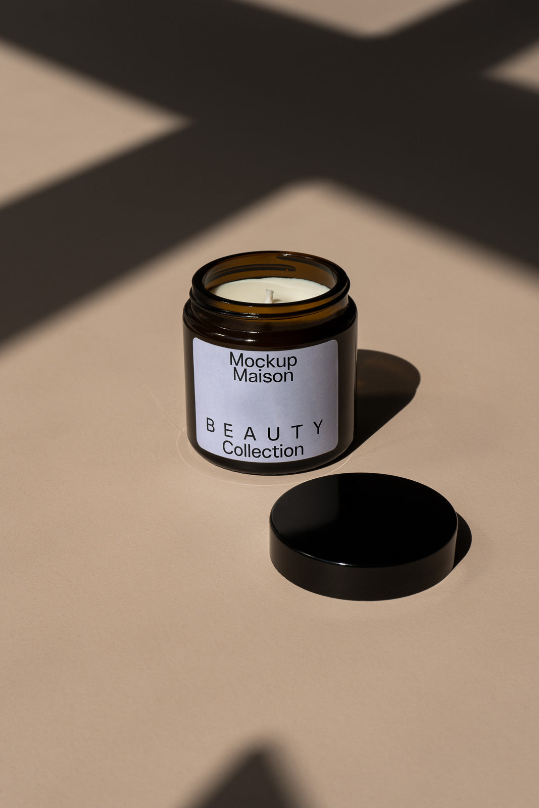 PSD mockup of an amber glass candle jar labeled "Mockup Maison Beauty Collection" with a black lid placed on a beige surface.