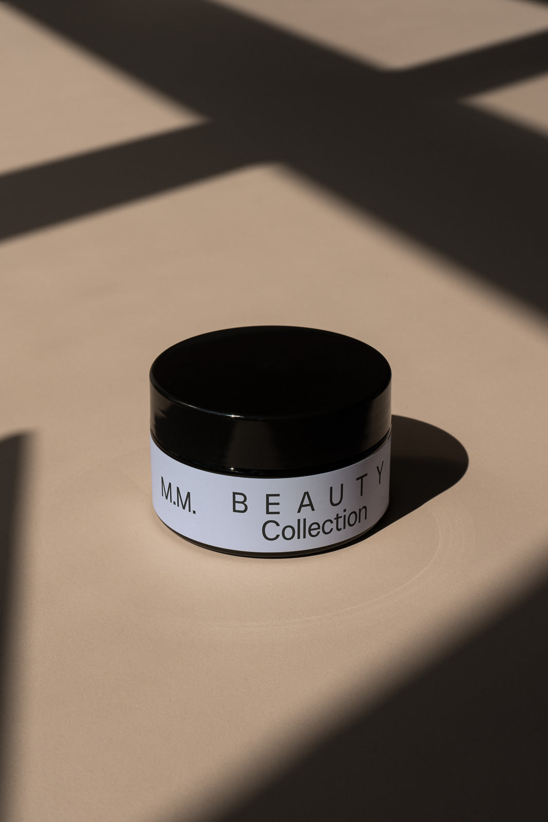 PSD mockup of a black cosmetic jar labeled "M.M. Beauty Collection" on a beige surface with dramatic shadows.