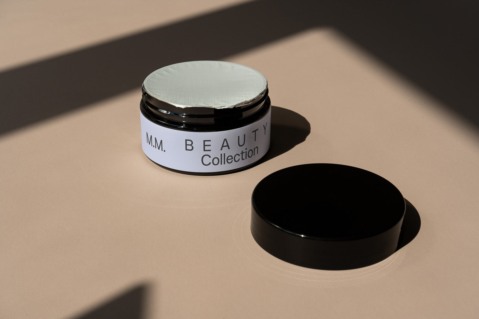 PSD mockup of a cosmetic jar labeled "M.M. Beauty Collection" with its lid off, placed on a beige surface with dramatic shadows.