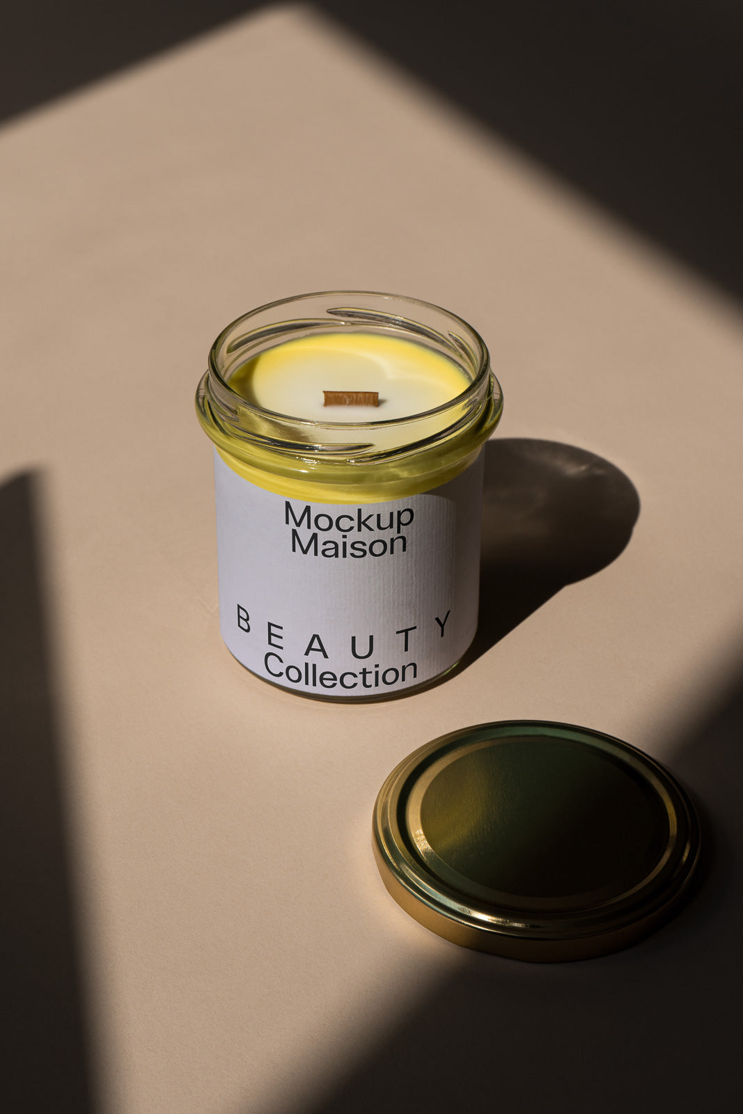 PSD mockup of a candle jar labeled "Mockup Maison Beauty Collection" with a metal lid on a beige surface, shadows adding depth to the composition.