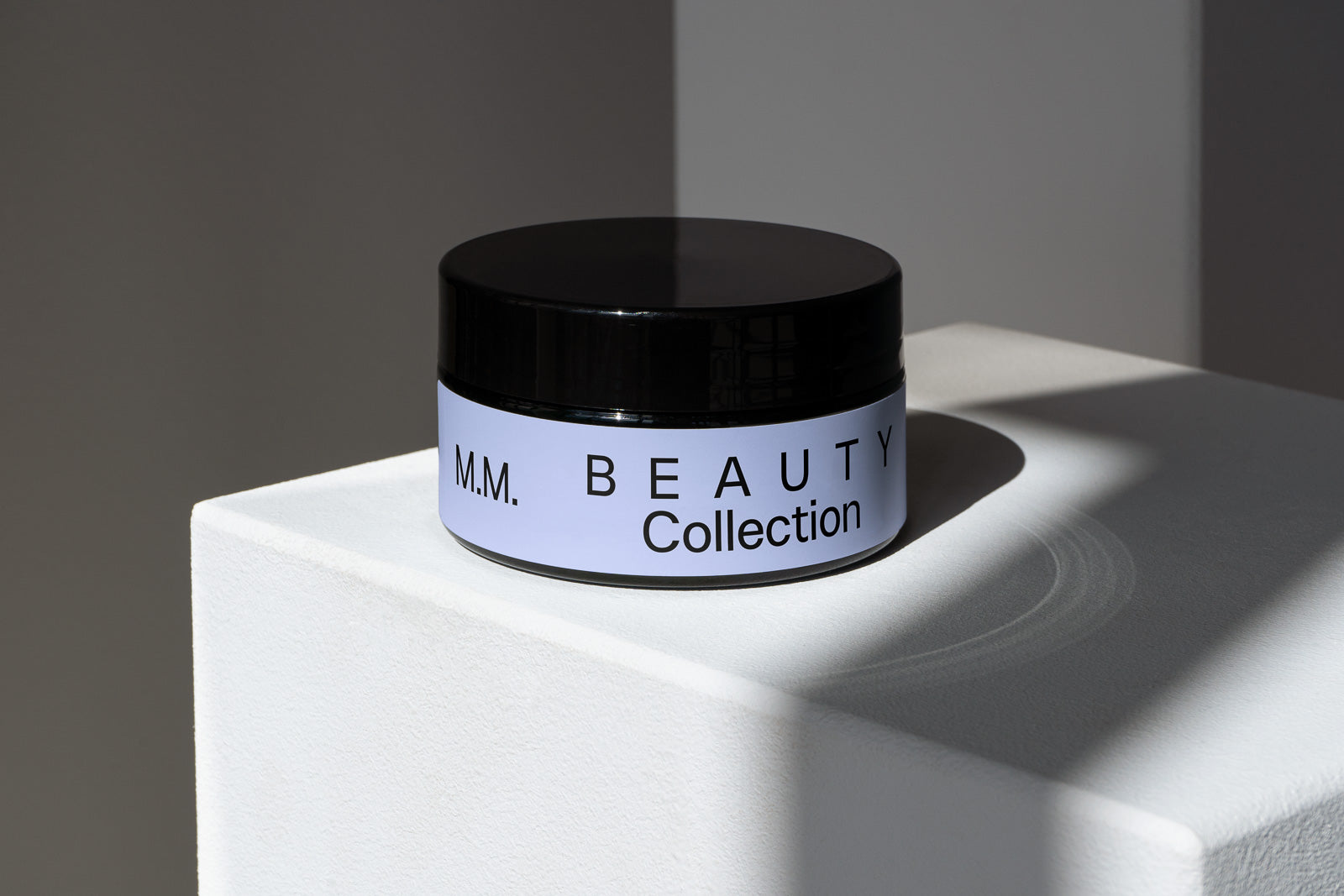PSD mockup of a cosmetic jar labeled "M.M. Beauty Collection" on a white block with soft shadows against a neutral background.