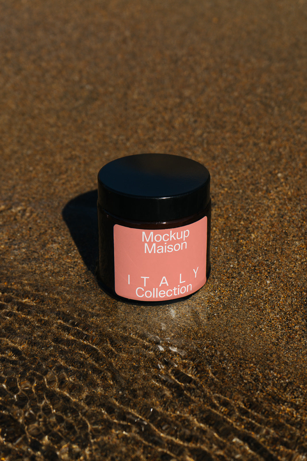 PSD mockup of a black jar with a coral label reading "Mockup Maison Italy Collection" placed on wet sand near rippling water.