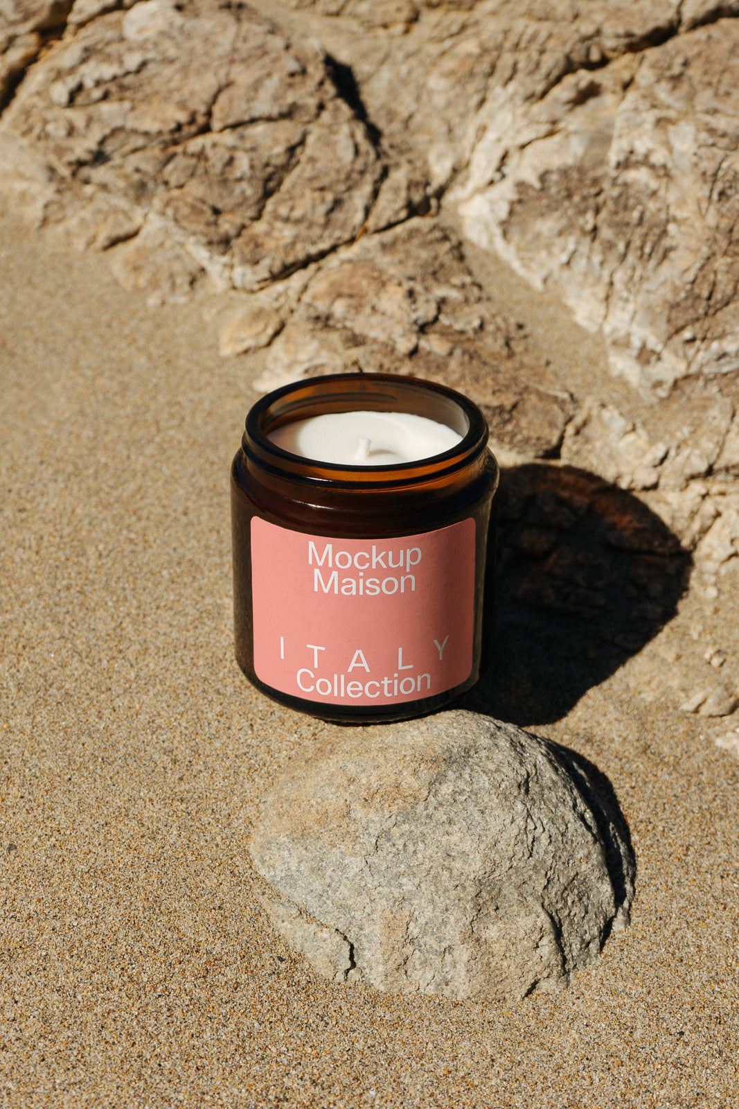 PSD mockup of a candle in a brown glass jar with a pink label reading "Mockup Maison Italy Collection," placed on sandy rocks.