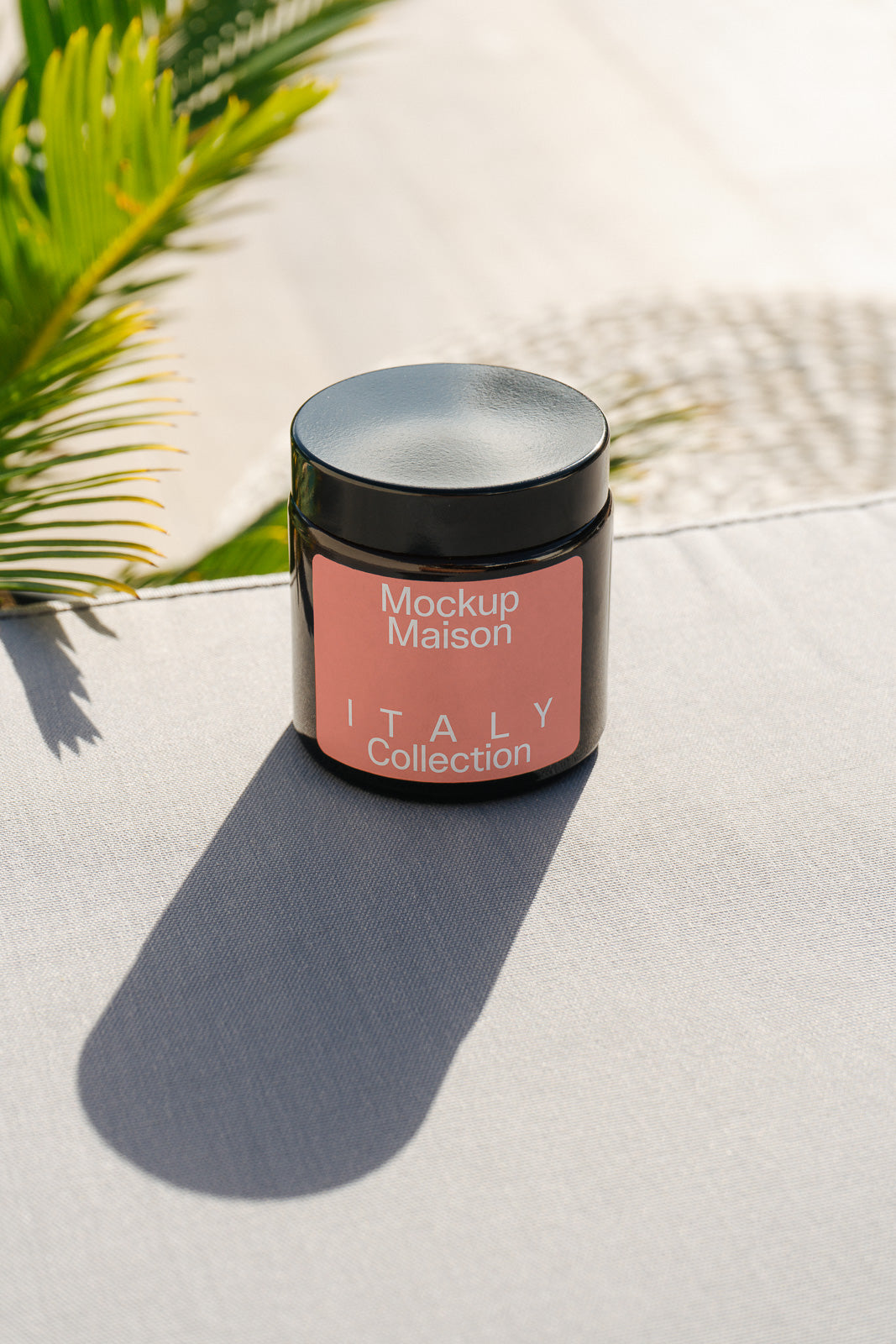 PSD mockup of a black jar with a pink label titled "Mockup Maison Italy Collection" placed on a surface in sunlight with a palm leaf shadow.