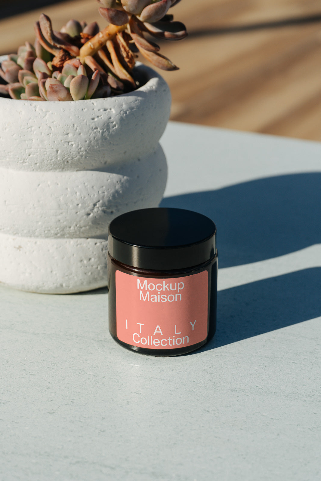 PSD mockup of a jar labeled "Mockup Maison Italy Collection" beside a textured white planter containing succulents on a smooth surface.