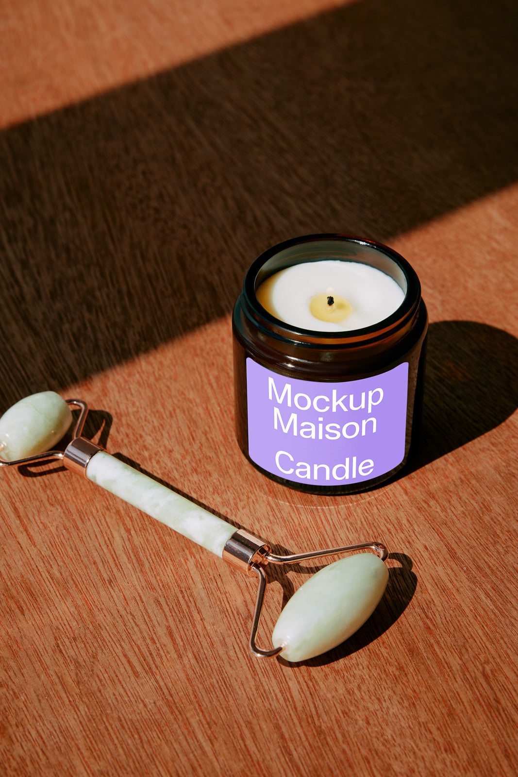 PSD mockup of a candle in a black jar labeled "Mockup Maison Candle" placed on a wooden surface alongside a jade facial roller.