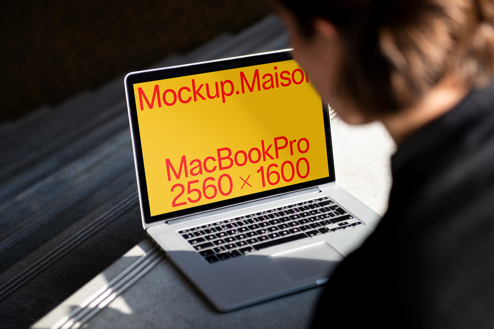 PSD mockup of a MacBook Pro with a yellow screen displaying the text "Mockup.Maison MacBookPro 2560 × 1600" in red, viewed by a person sitting on steps.