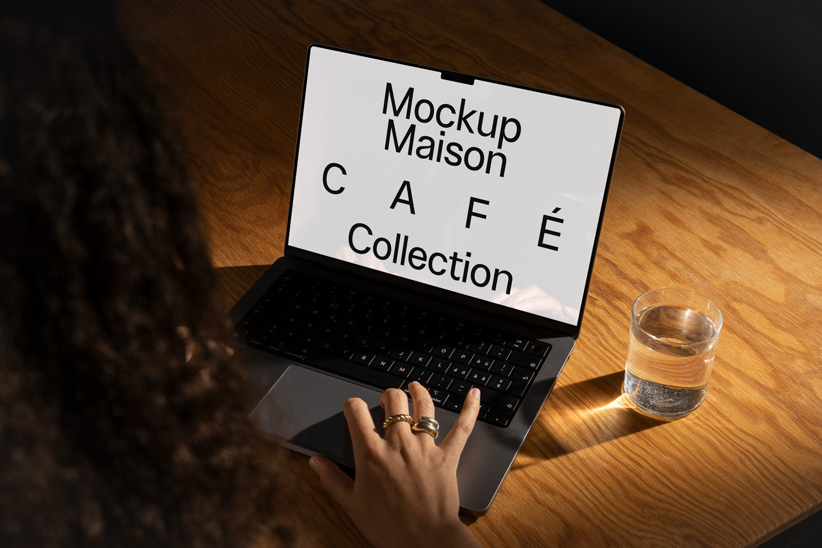 PSD mockup of a laptop displaying "Mockup Maison CAFÉ Collection" on a wooden table, with a hand poised over the keyboard and a glass of water nearby.