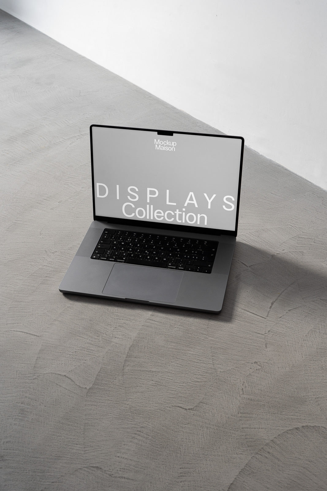 PSD mockup of an open laptop on a concrete floor, displaying a screen with the text "Mockup Maison Displays Collection" in a minimalist setting.