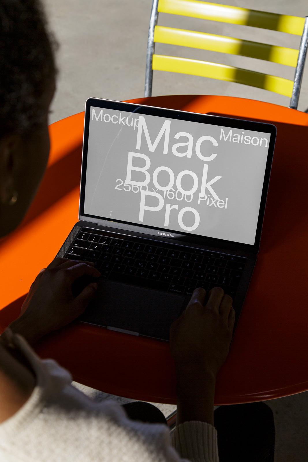 PSD mockup of a MacBook Pro on a bright orange table, displaying design text on the screen, with hands typing on the keyboard.