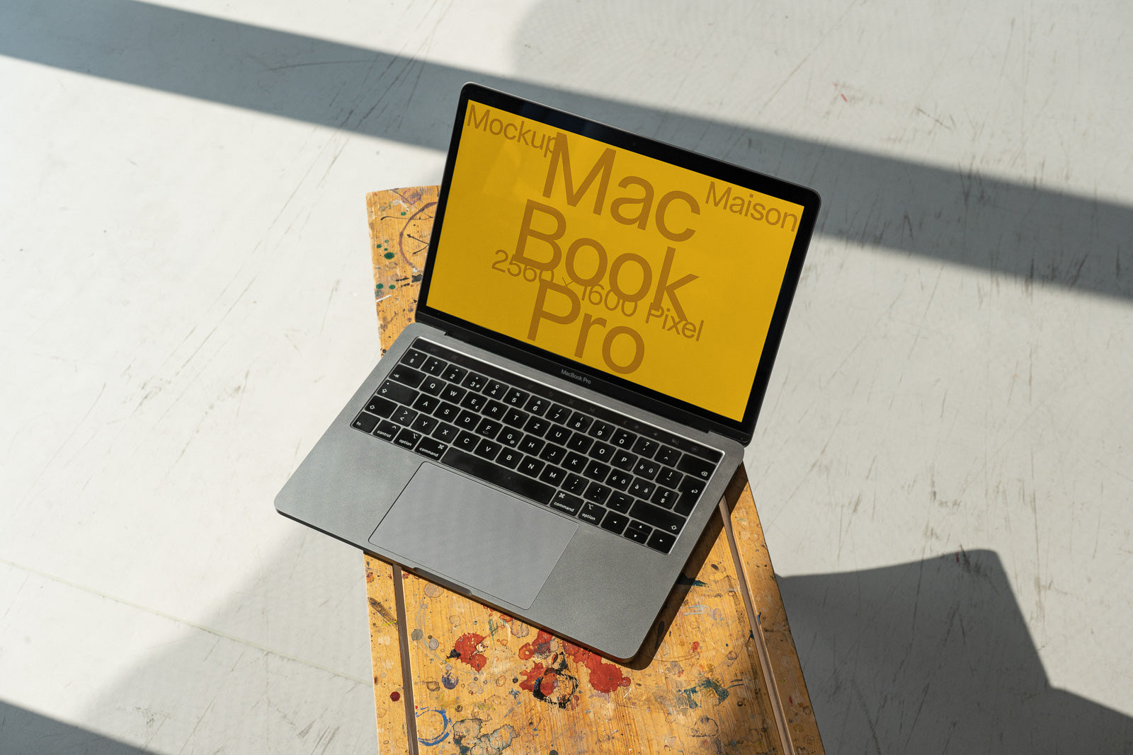 PSD mockup of a MacBook Pro on a wooden stool with a yellow screen displaying text. Sunlight casts shadows on a white floor.