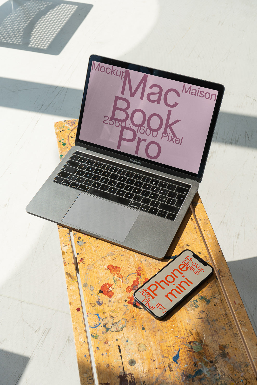 PSD mockup of a MacBook Pro and iPhone mini on a wooden table covered with colorful paint splatters, set in bright natural light.