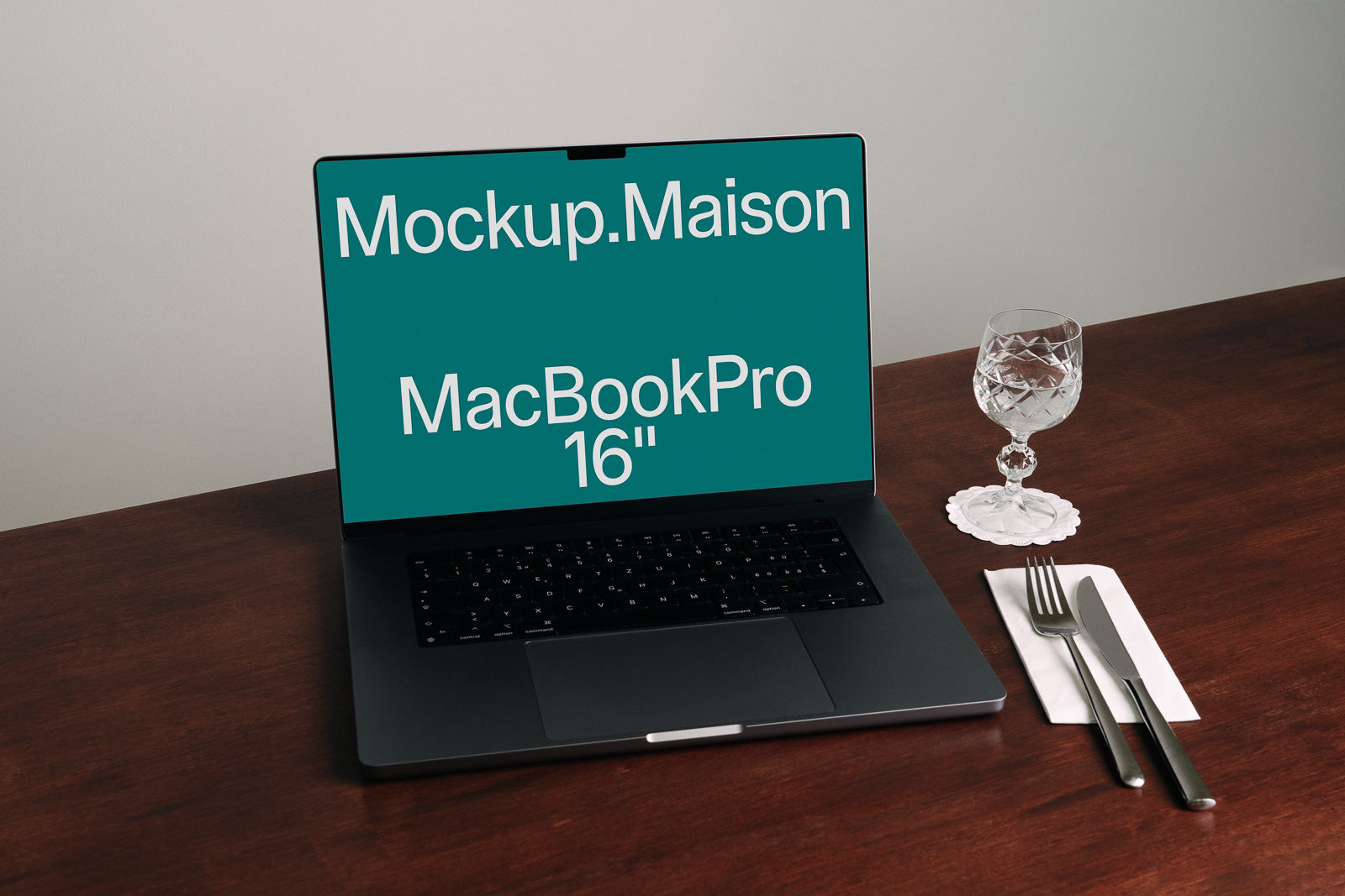 PSD mockup of a MacBook Pro 16-inch on a wooden table with cutlery and a crystal glass nearby.