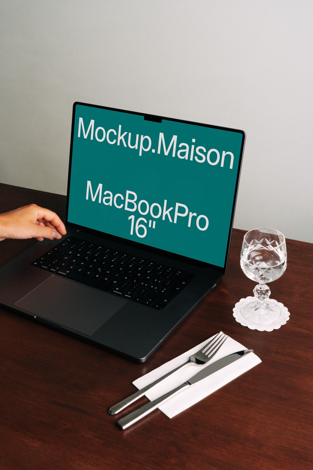 PSD mockup of a 16" MacBook Pro on a wooden table with a hand adjusting its screen. Nearby, a glass and cutlery are placed on a napkin.
