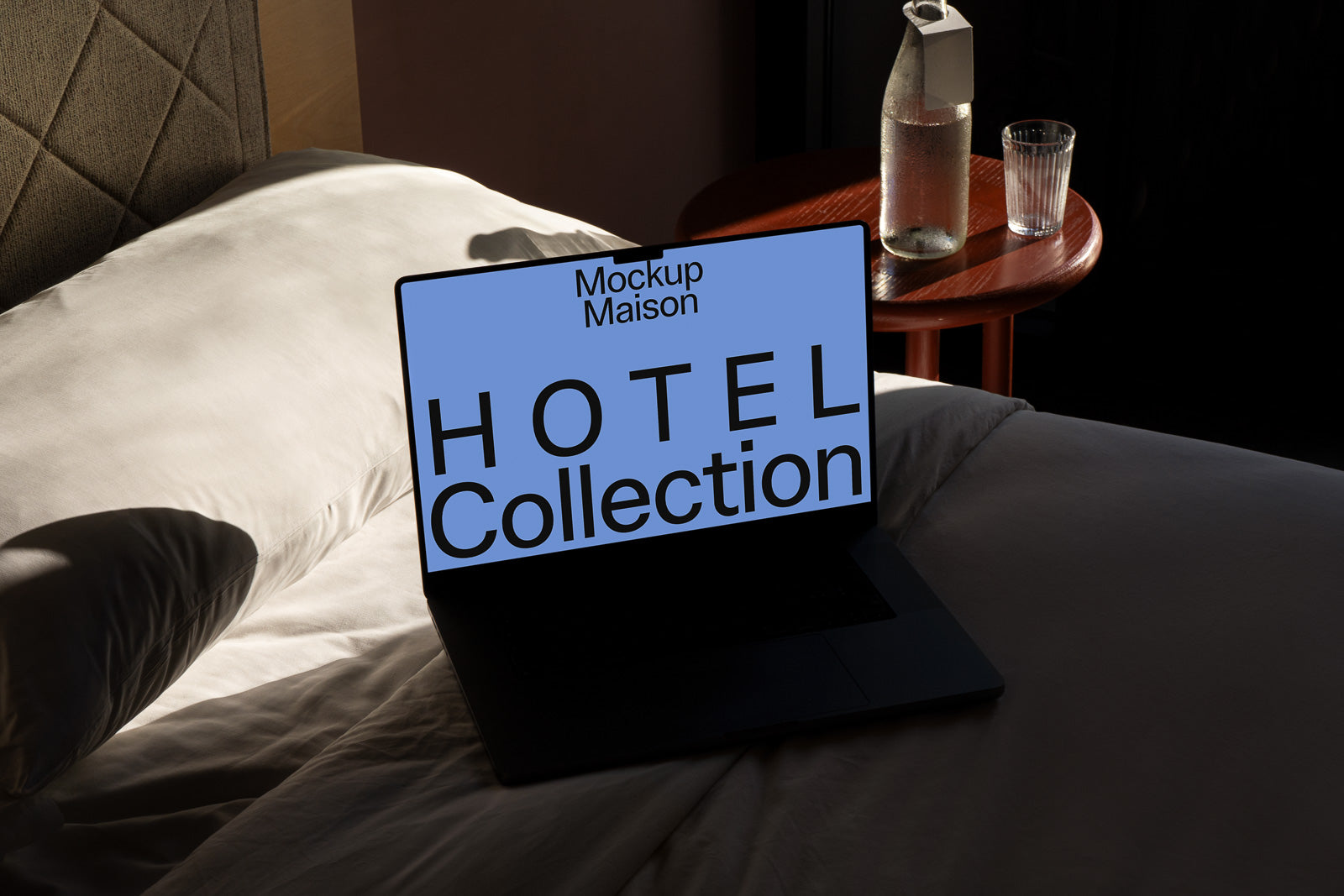 PSD mockup of a laptop on a bed displaying "Hotel Collection" text on the screen, with a glass bottle on a nearby wooden table.