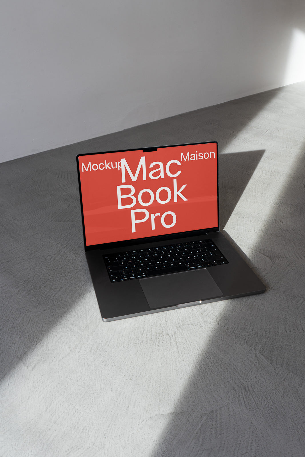 PSD mockup of a MacBook Pro with a red screen displaying the text "MacBook Pro" and "Maison Mockup," placed on a gray textured surface with sunlight shadows.