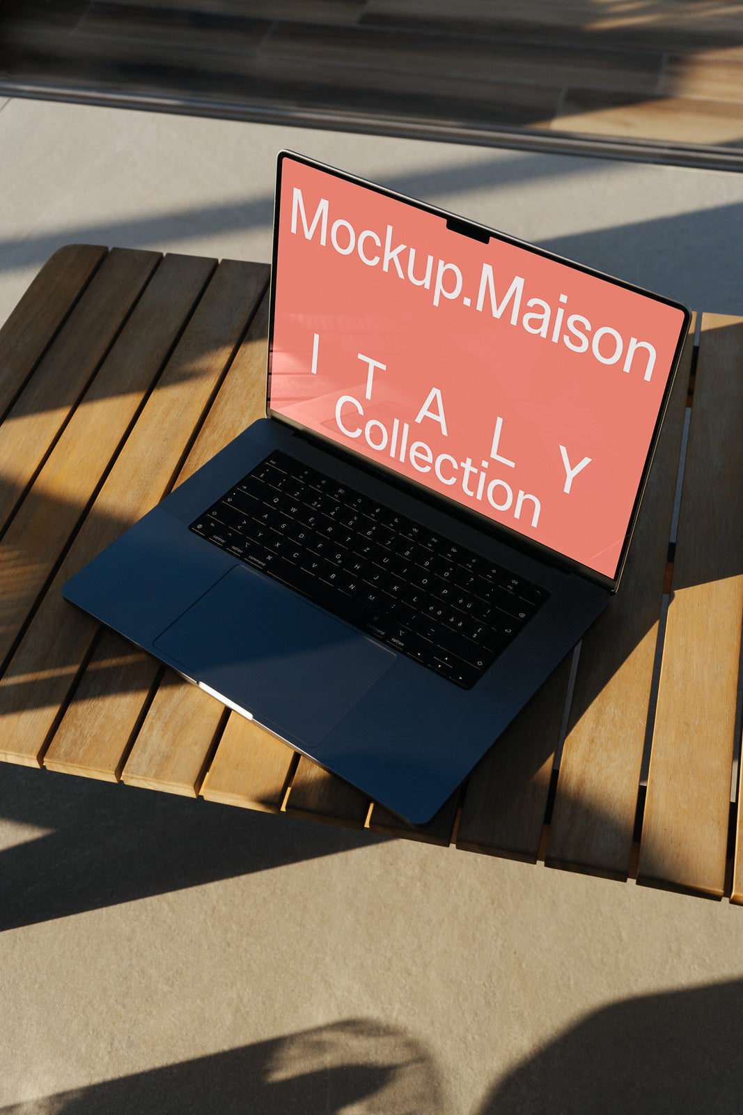 PSD mockup of a laptop on a wooden table displaying a pink screen with the text "Mockup.Maison Italy Collection" in a sunlit setting.