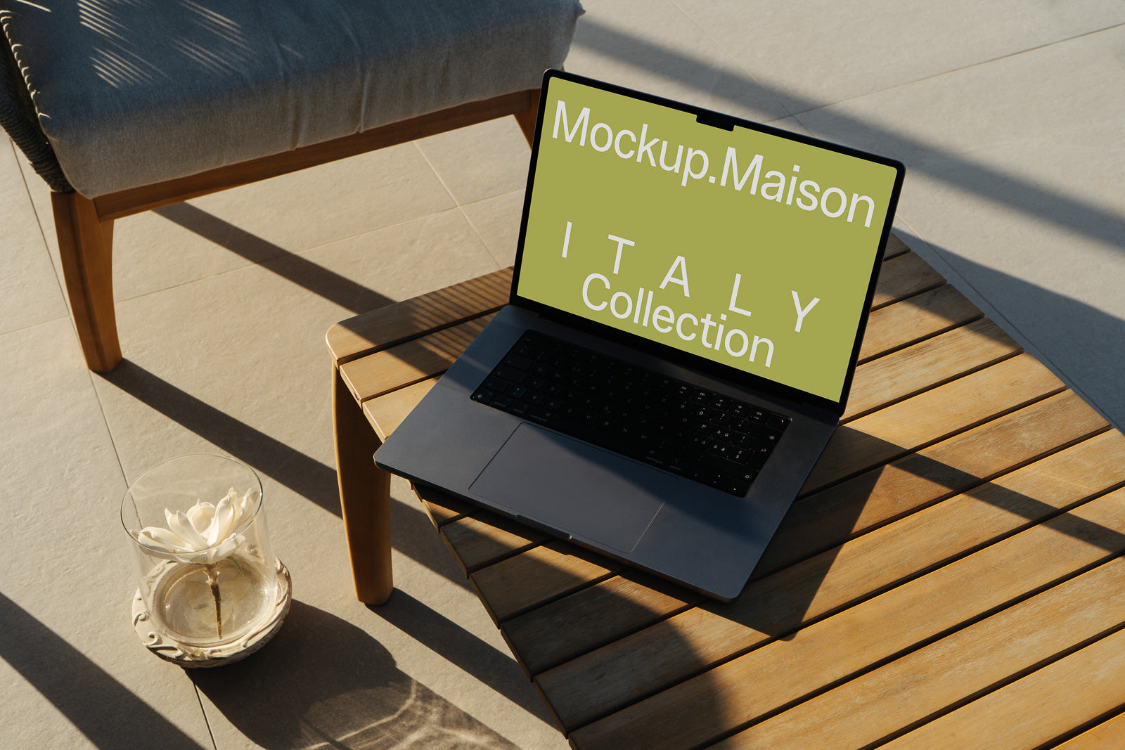 PSD mockup of a laptop on a wooden table with a green screen displaying "Mockup.Maison ITALY Collection" beside a flower in a glass vase.