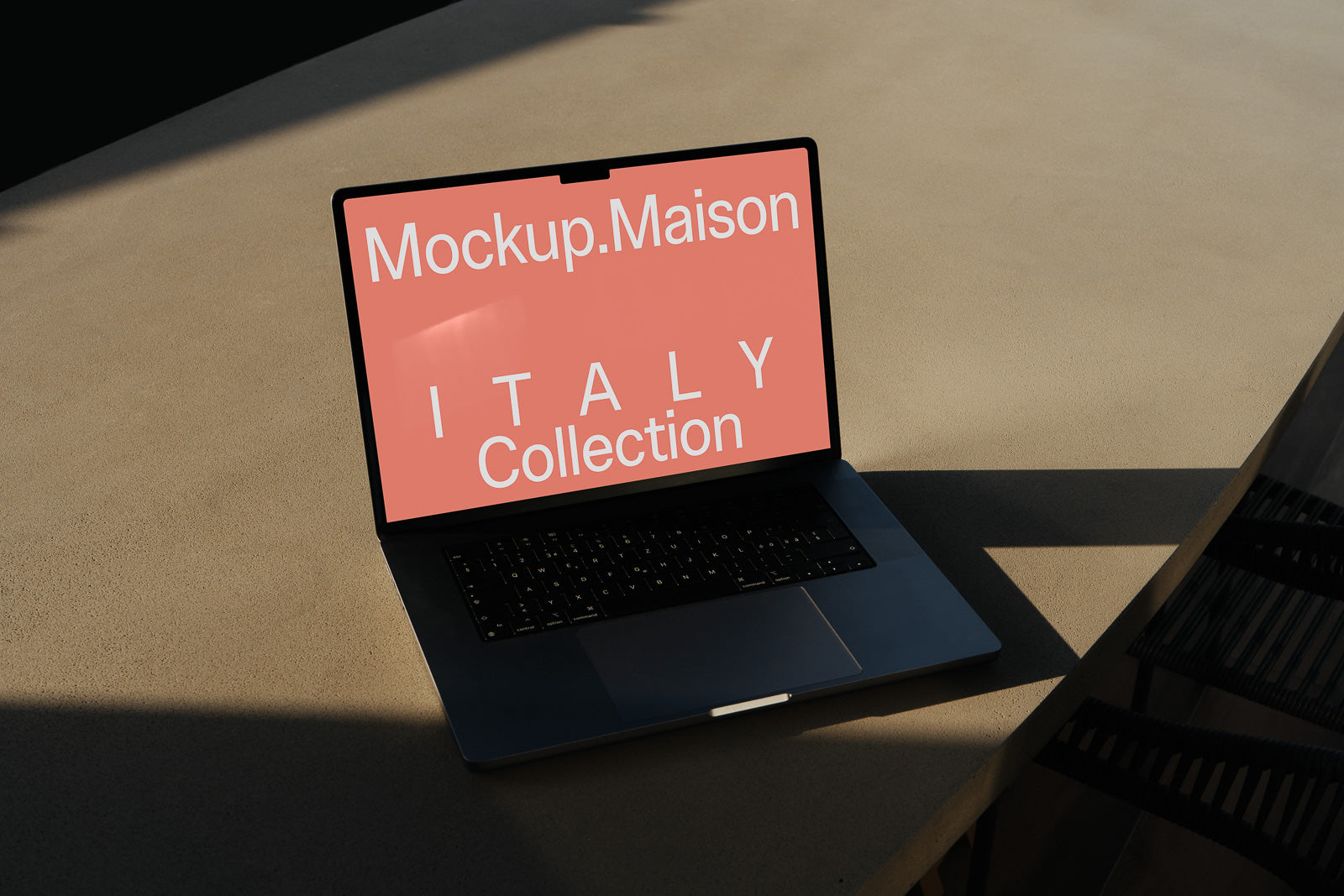 PSD mockup of a sleek laptop displaying "Mockup.Maison ITALY Collection" on a coral background, placed on a light-colored table with shadow.