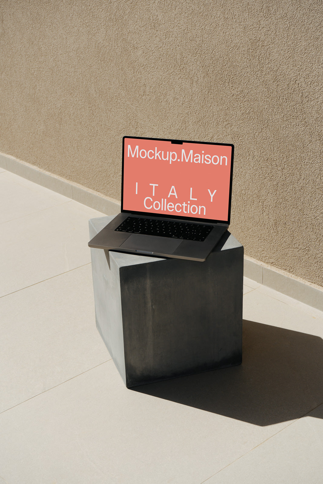PSD mockup of a laptop displaying "Mockup.Maison ITALY Collection" on a textured backdrop, set atop a concrete block in an outdoor setting.