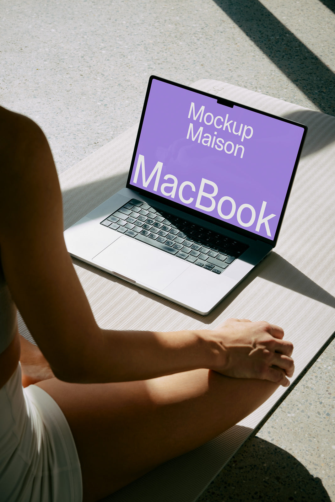 PSD mockup of a MacBook displaying "Mockup Maison" on a purple screen, placed on a textured surface next to a seated person in workout attire.
