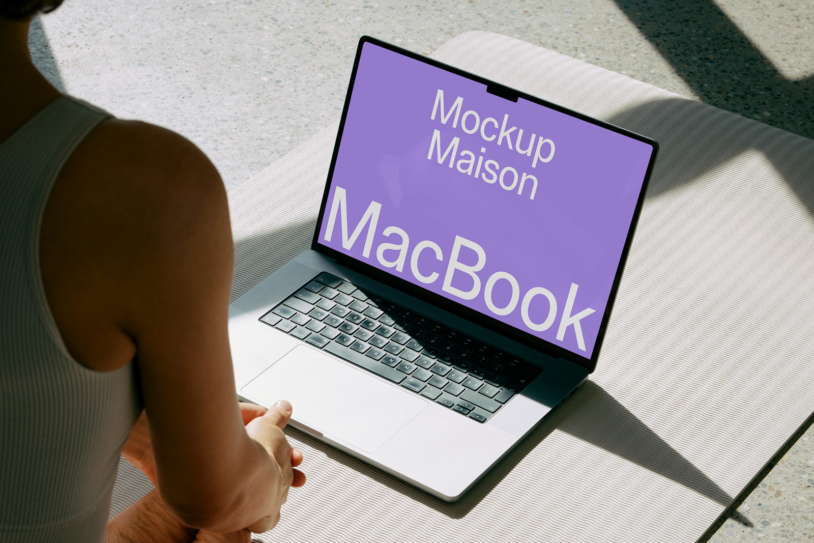 PSD mockup of an open MacBook on a light surface, displaying a purple screen with text "Mockup Maison MacBook," viewed from behind a seated person.