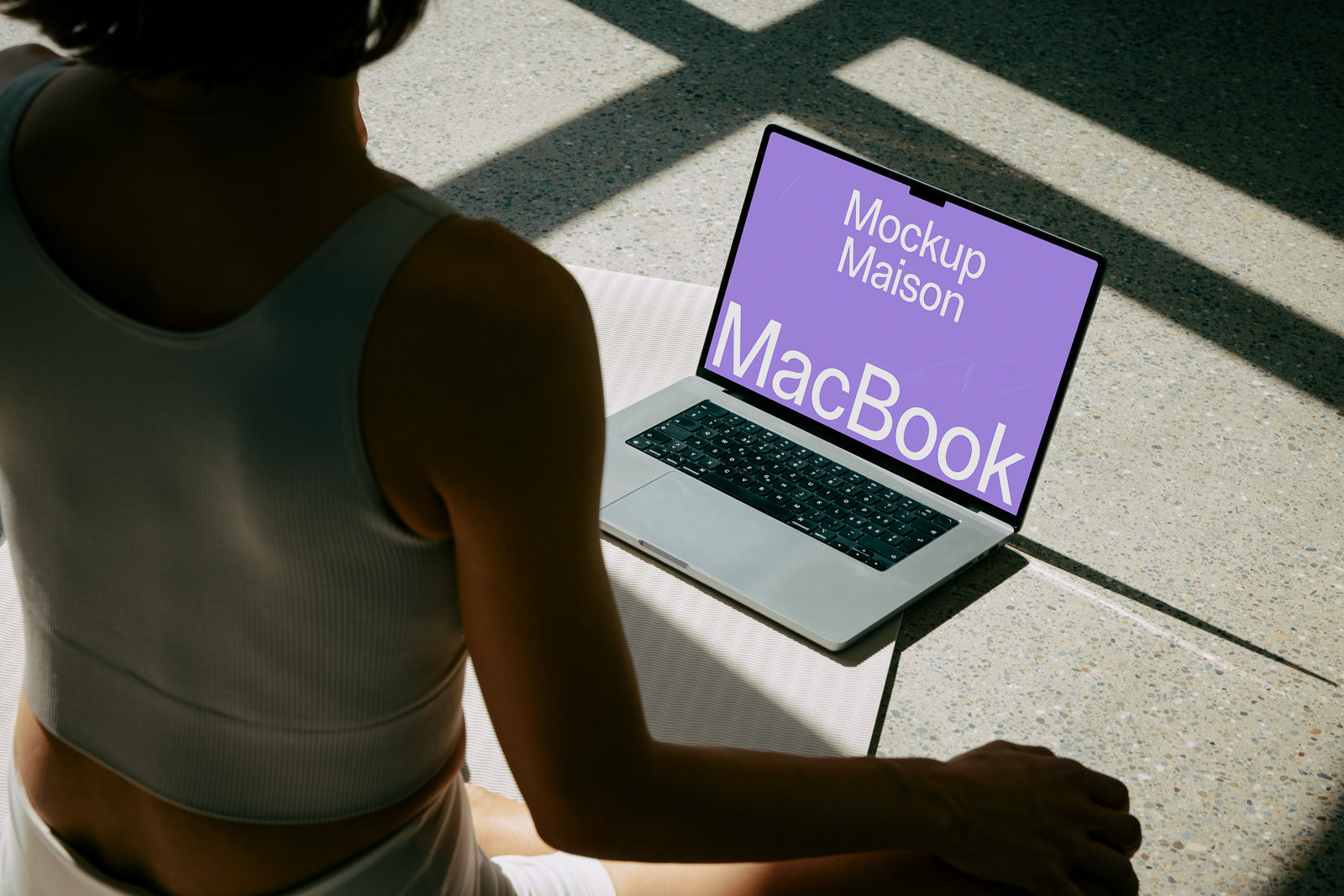 PSD mockup of an open MacBook placed on a textured floor with a woman sitting nearby, displaying a screen with "Mockup Maison" in prominent text.
