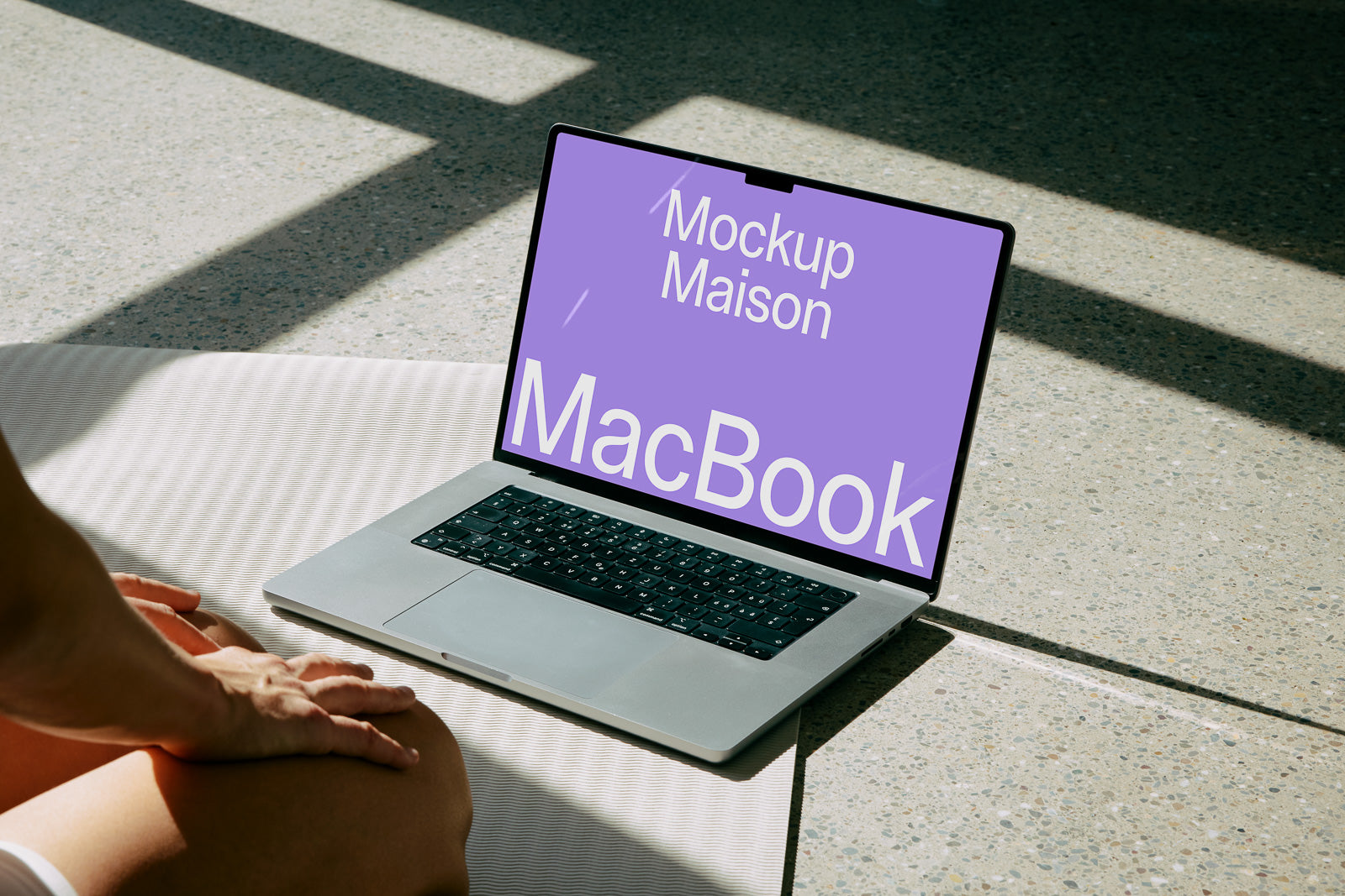 PSD mockup of a MacBook displaying text "Mockup Maison MacBook" on the screen, placed on a light-colored surface with person sitting nearby.
