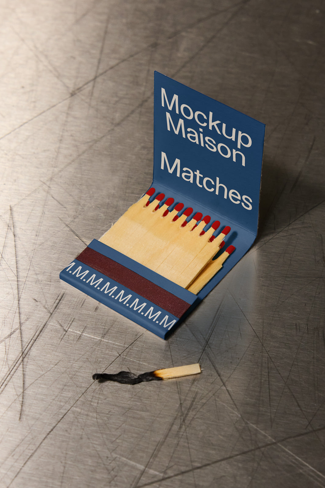 PSD mockup of an open matchbook with the text "Mockup Maison Matches" on the cover, placed on a textured metallic surface. A burnt match lies nearby.