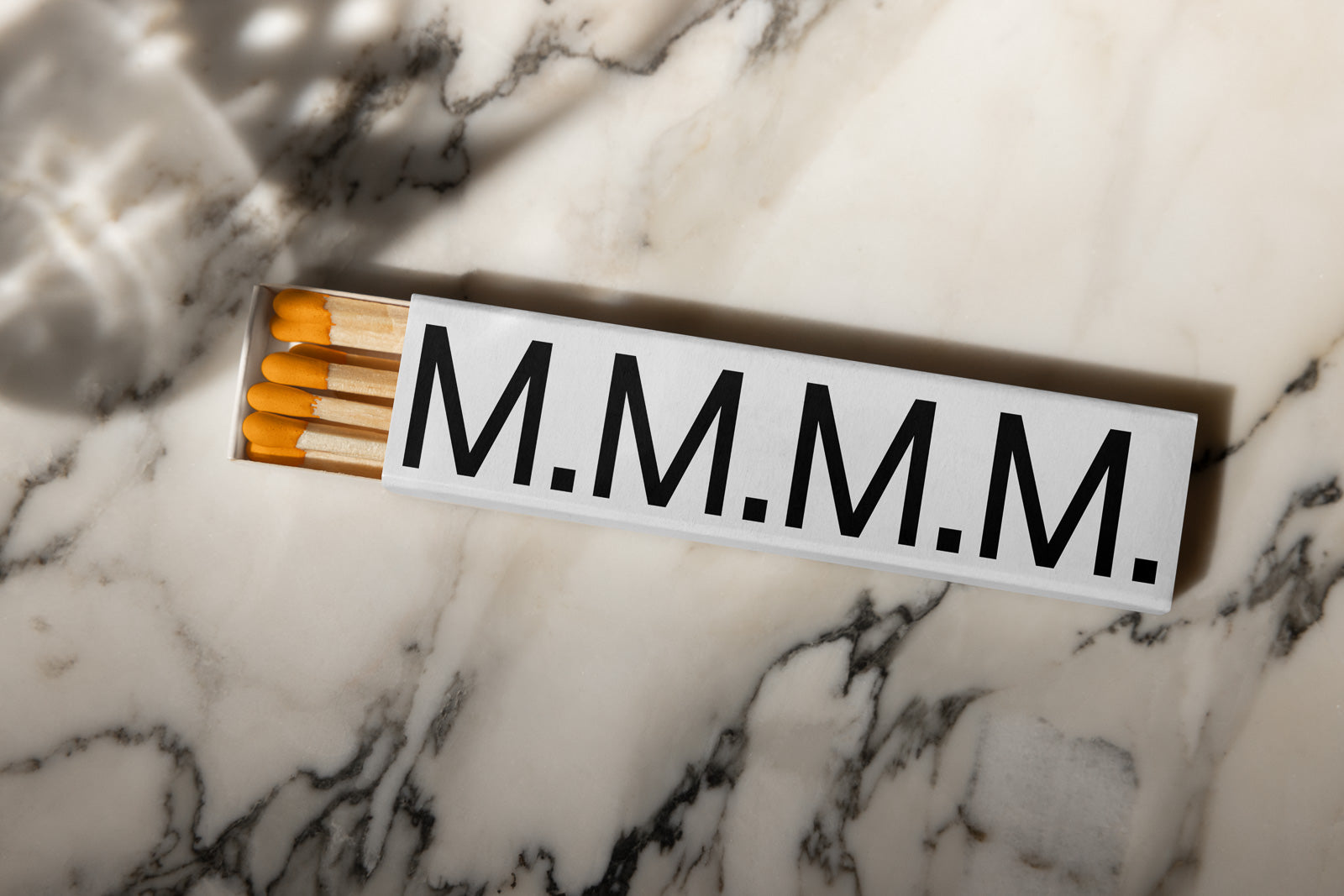 PSD mockup of a matchbox with bold letters "M.M.M.M." on a marble surface, open to reveal several matches with orange tips.