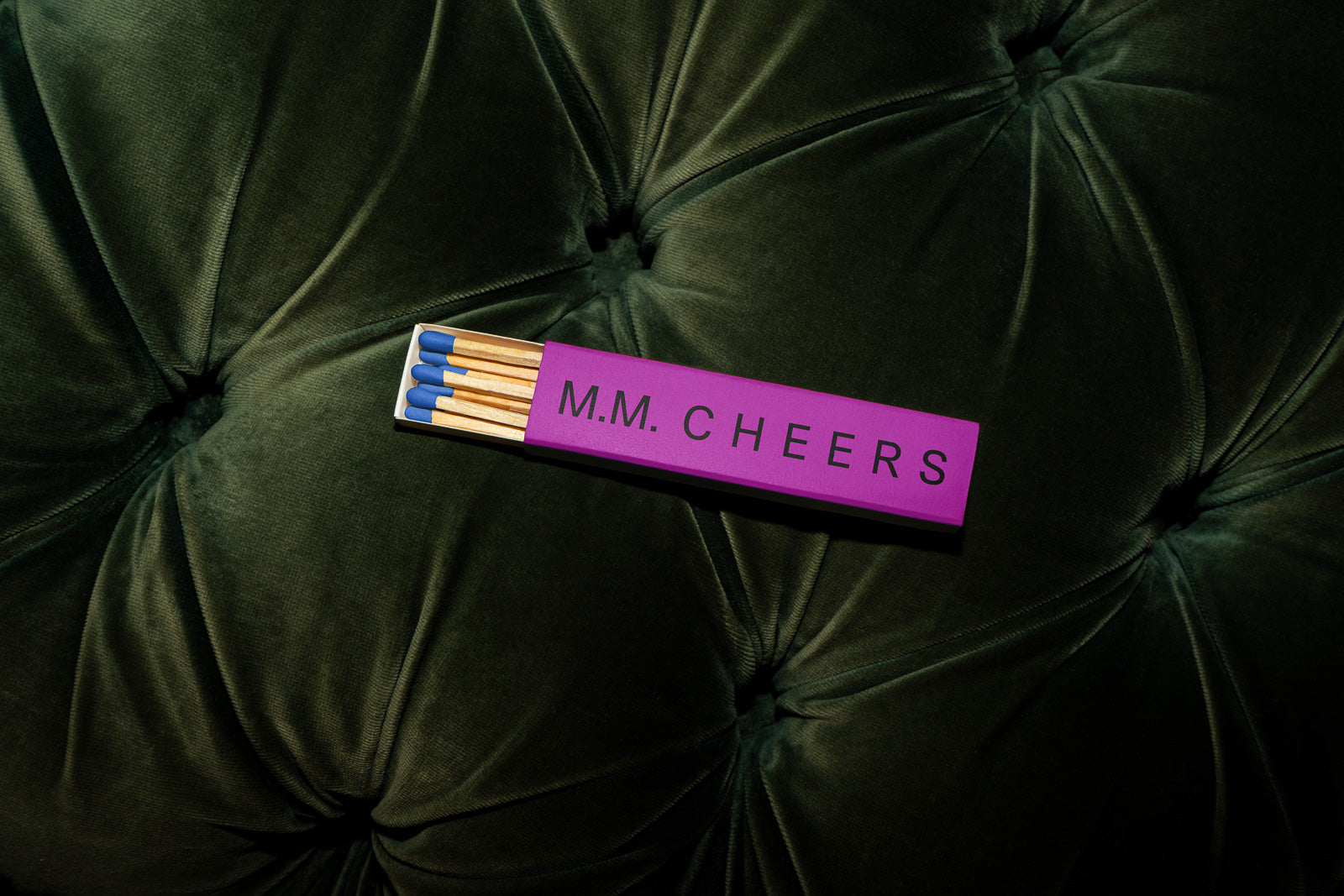 PSD mockup of a matchbox with "M.M. CHEERS" written on it, placed on a dark green tufted fabric surface.