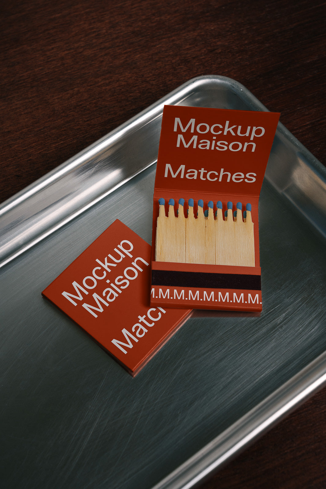 PSD mockup of two matchbooks on a metal tray, one open displaying matches with red and blue tips, both featuring white text on a red cover.