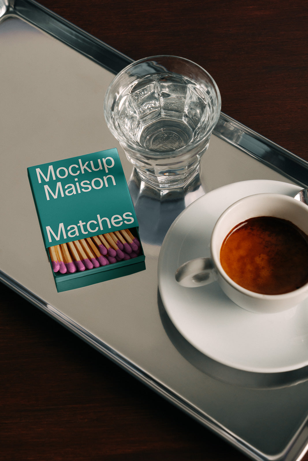 PSD mockup of a matchbox labeled "Mockup Maison Matches" on a silver tray beside a glass of water and a cup of espresso on a wooden table.