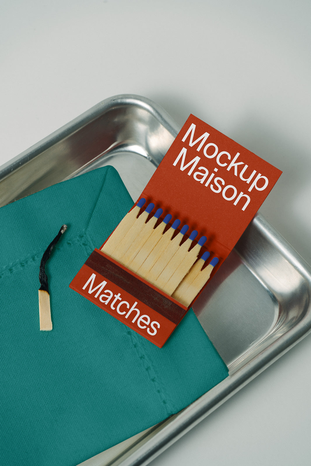 PSD mockup of a matchbook labeled "Mockup Maison" with blue-tipped matches, placed on a metal tray beside a burnt matchstick on a teal fabric.