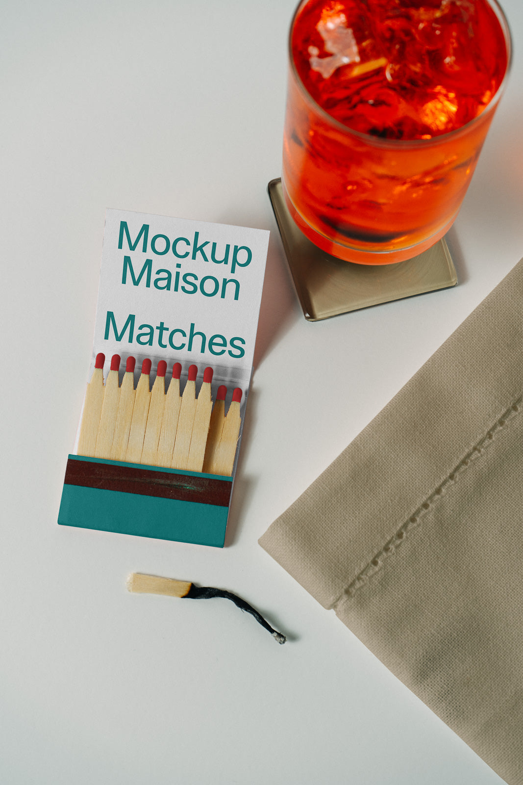 PSD mockup of an open matchbook labeled "Mockup Maison Matches" beside a red drink in a glass on a coaster, with a folded napkin nearby.