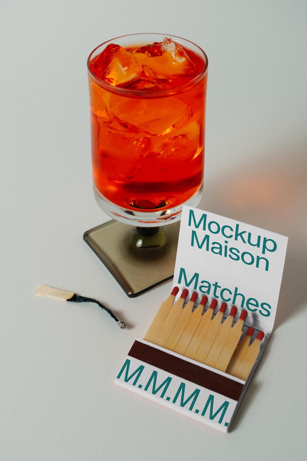 PSD mockup of a glass with an orange drink and ice cubes beside an open matchbook labeled "Mockup Maison Matches."
