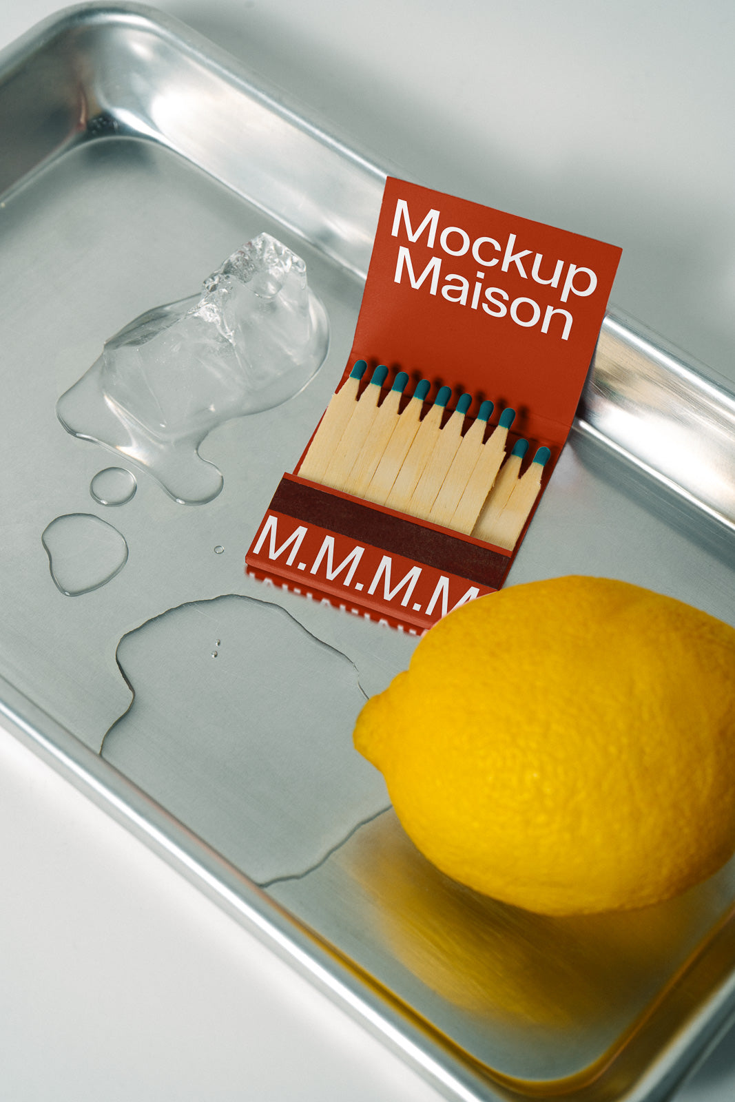 PSD mockup of a matchbook with "Mockup Maison" text, placed on a metal tray with a lemon, ice cube, and water droplets.