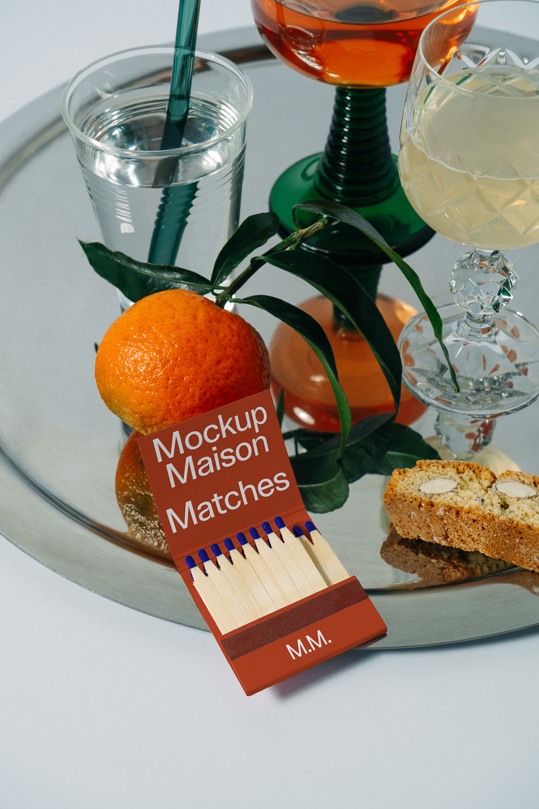 PSD mockup of a matchbook with "Mockup Maison Matches" text, set on a tray with drinks, an orange, and biscuit, creating an elegant scene.