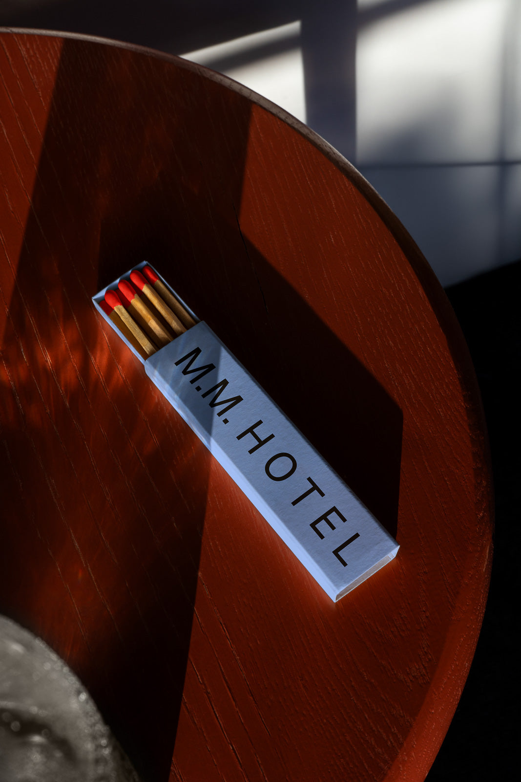 PSD mockup of a matchbox labeled "M.M. Hotel" resting on a round wooden table, with shadows cast across the surface.