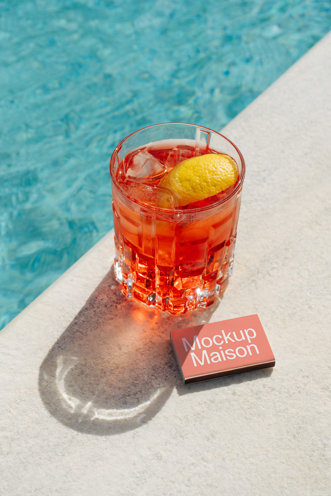 PSD mockup of a vibrant cocktail in a glass with ice and a lemon wedge by the poolside, accompanied by a small 'Mockup Maison' card on the edge.