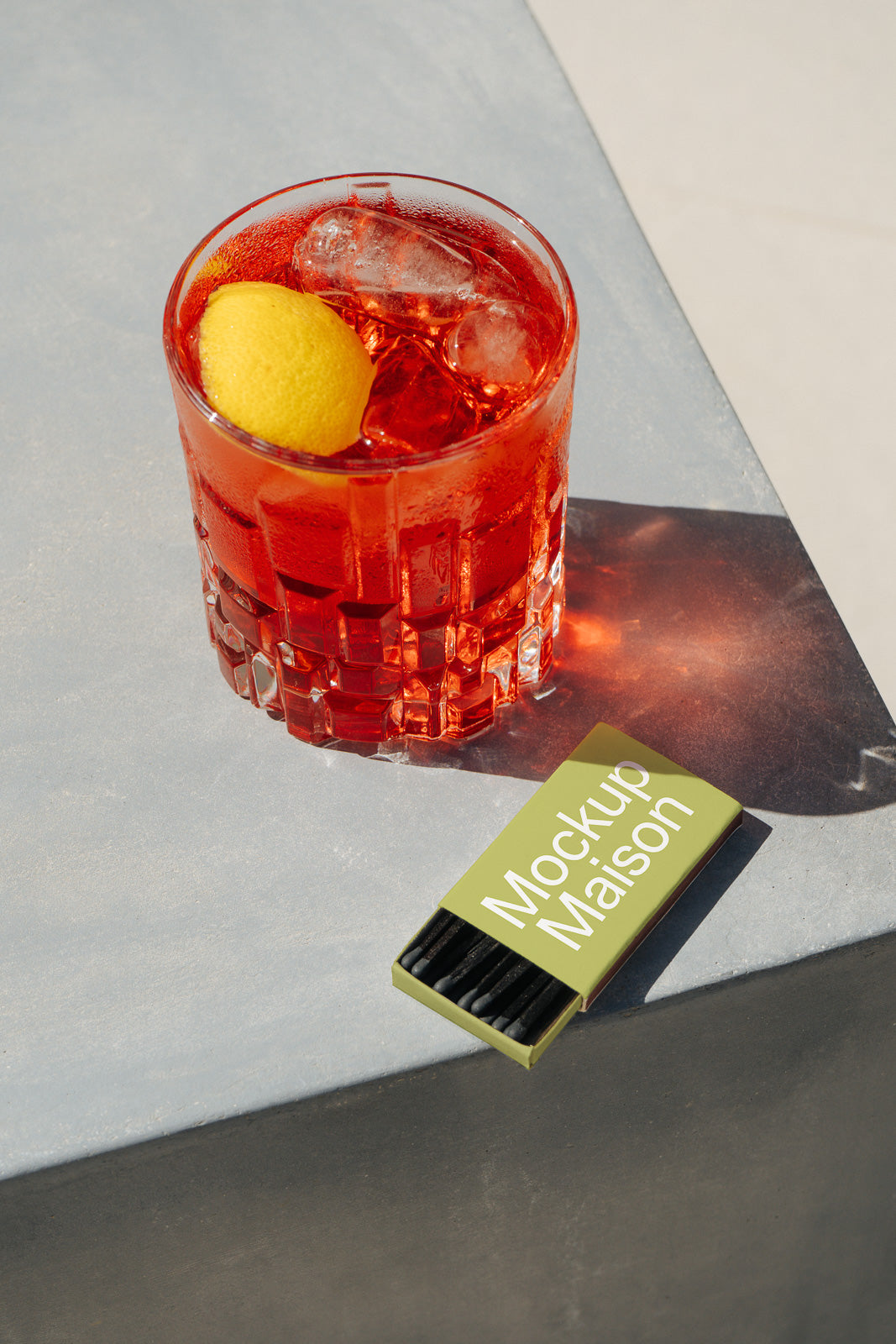 PSD mockup of a clear glass with a red cocktail, ice cubes, and a lemon peel garnish on a table beside a box of matches labeled "Mockup Maison."