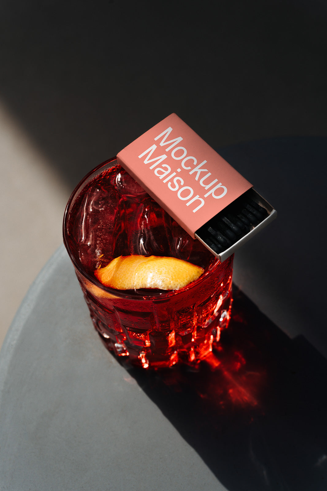PSD mockup of a cocktail glass with a red beverage and an orange peel garnish, with a matchbox labeled "Mockup Maison" resting on the rim.
