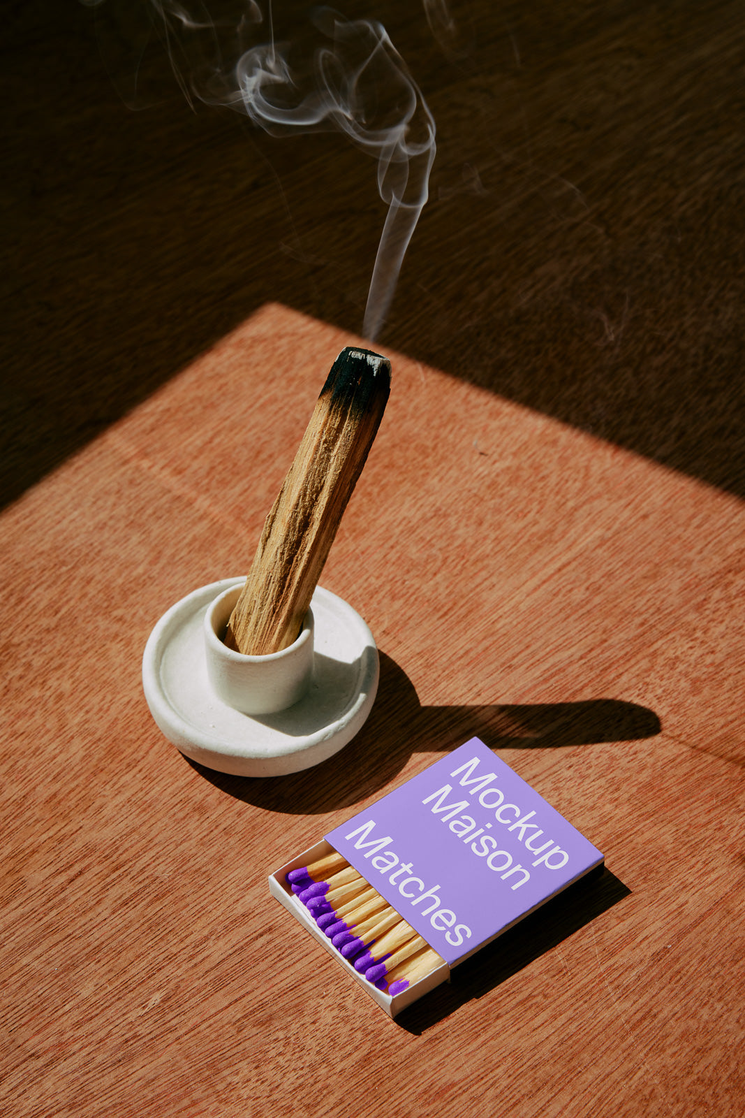 PSD mockup of a lit incense stick in a holder next to a matchbox with purple match tips, on a wooden surface.