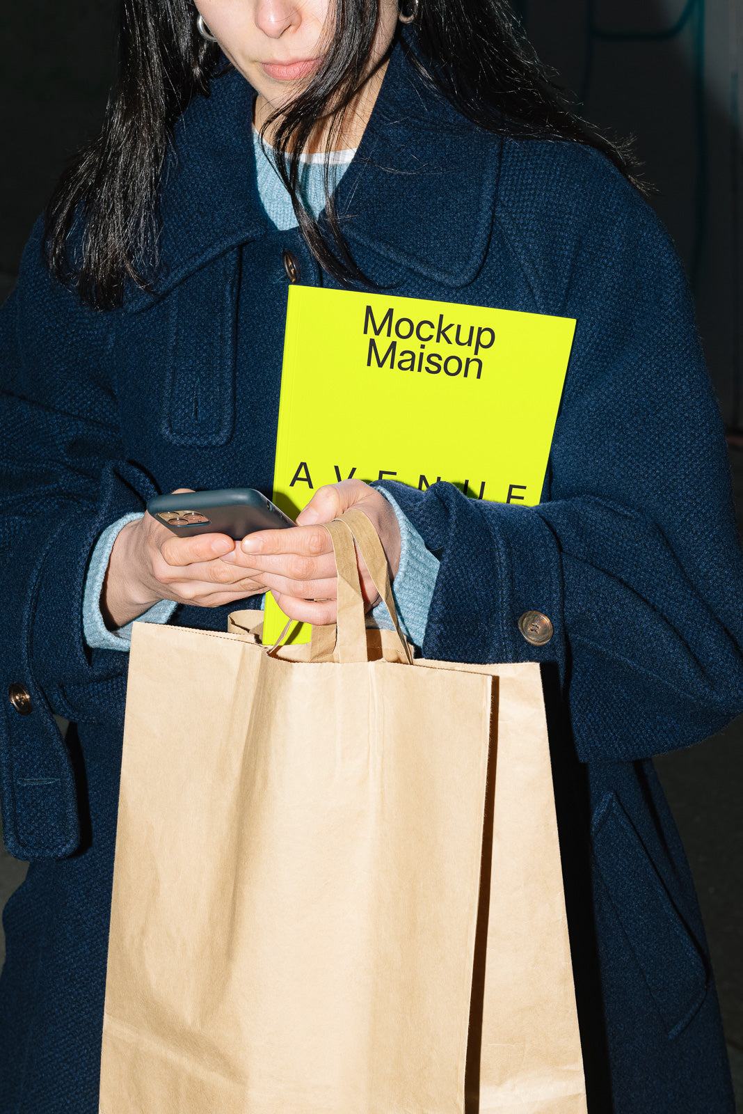 PSD mockup of a person holding a bright yellow booklet titled "Mockup Maison Avenue" along with a paper shopping bag, checking their phone.
